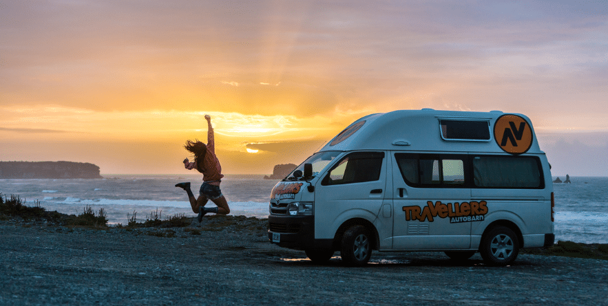 Person jumping next to campervan with sunset view over ocean, New Zealand
